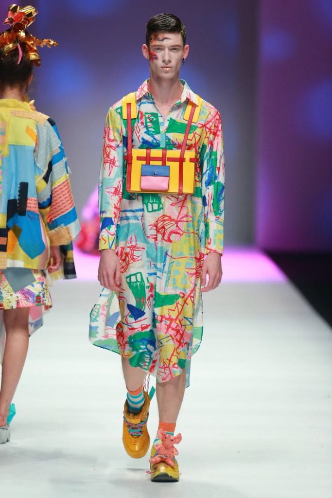 Model walking the runway at Shanghai Fashion Week wearing a colorful abstract-patterned outfit with bold accessories, showcasing Angel Chen's vibrant Spring/Summer 2015 collection.