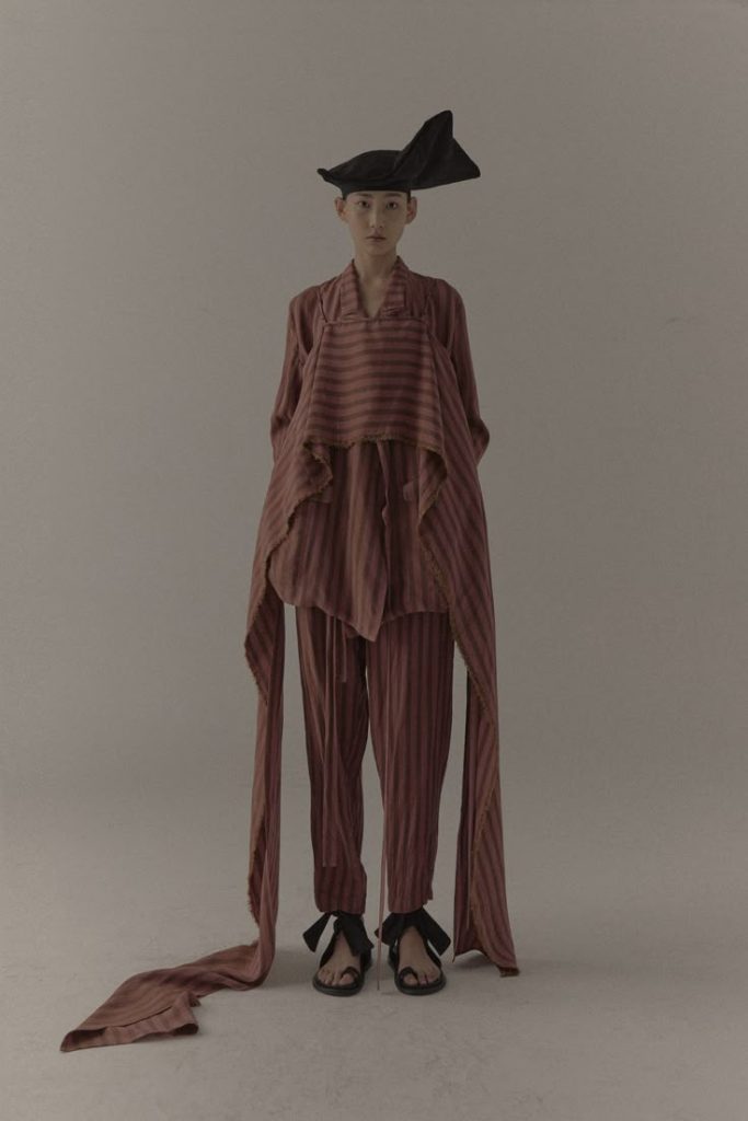 Model in striped, layered ensemble with asymmetric draping from Uma Wang Spring 2021 Ready-to-Wear collection, styled with a sculptural headpiece.